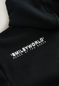 Sort hættetrøje med hvidt trykt logo, der lyder "SMILEYWORLD" og "BAKER BY TED BAKER," med en tekstureret finish omkring trykket.
