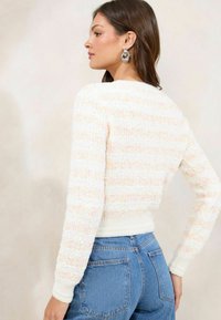 White textured sweater with pastel stripes, fitted design, and ribbed cuffs, paired with high-waisted blue jeans. Seam details are visible.