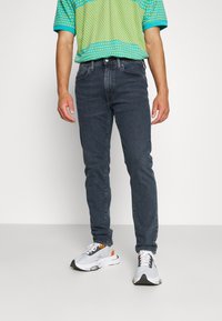 Jeans de mezclilla azul oscuro con corte slim, combinados con una camisa de patrón vibrante en verde y amarillo y zapatillas deportivas grises.