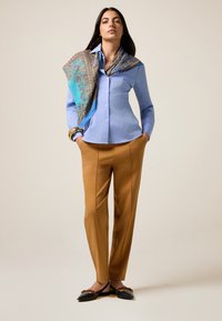 Chemise bleu clair à boutons, pantalon beige, écharpe à motifs bleu et marron, et ballerines noires avec un détail décoratif, mains dans les poches.