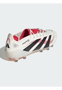 Weiße und schwarze Fußballschuhe mit roten Akzenten. Texturiertes Obermaterial, sechs stollenförmige Außensohle für Traktion und "Predator"-Branding auf der Seite.