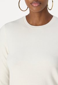 Krämfärgad stickad tröja med ribbat runt halsen, slät textur och långa ärmar. Guldringörhängen delvis synliga.