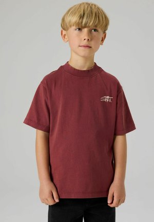 Camiseta de manga corta color marrón hecha de algodón. Presenta un cuello redondo y un pequeño logo blanco en el pecho. Se lleva con pantalones negros.