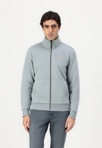 Džepar sweatshirt od svijetlosive tkanine, s visokim ovratnikom, rebrastim manžetama i prednjim džepom; mali zlatni logo na prsima.