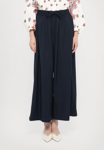 Navy wide-leg trousers made of soft fabric with an elastic waistband and drawstring; worn with pointed-toe beige shoes.