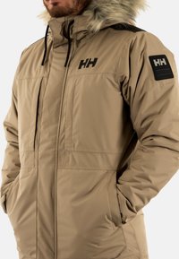 Parka imbottita beige con cappuccio foderato in pelliccia, zip frontale, due tasche laterali e un patch del logo nero sulla spalla sinistra.