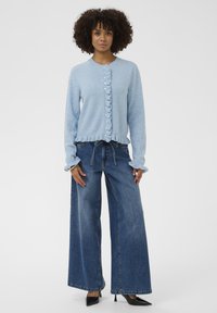 Világoskék kardigán fodros széllel és gombos elülső résszel, párosítva széles szárú sötét farmerral és fekete magassarkú cipőkkel.