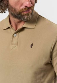 Homme barbu portant un polo beige avec col boutonné et petit logo brodé sombre sur la poitrine gauche.