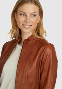 Braune Lederjacke mit hohem Kragen, Reißverschluss vorne und Nahtdetails. Über einem hellen Hemd getragen, vor einem neutralen Hintergrund.