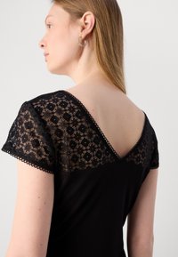Robe noire avec des manches en dentelle au motif géométrique, décolleté en V au dos et silhouette fluide. Texture subtile et coupe élégante.