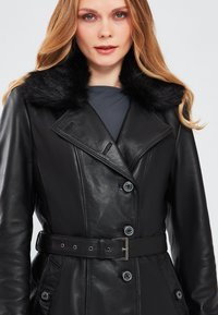 Cappotto in pelle nera con colletto in pelliccia sintetica, bottoni doppi, vita cintata e tasche laterali. Texture liscia, vestibilità su misura.