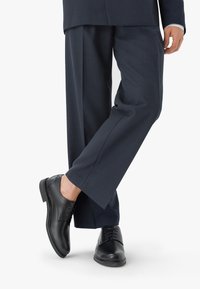 Czarne skórzane buty dressowe o gładkim wykończeniu, zapinane na sznurowadła, z okrągłym noskiem, w zestawieniu z ciemnymi spodniami w prążki.