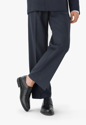 Czarne skórzane buty dressowe o gładkim wykończeniu, zapinane na sznurowadła, z okrągłym noskiem, w zestawieniu z ciemnymi spodniami w prążki.