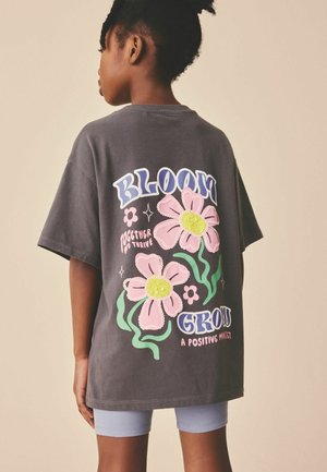 Copil purtând un tricou supradimensionat gri închis cu flori roz și textul „Bloom Grow A Positive Mindset” pe spate, asortat cu pantaloni scurți albastru deschis.