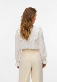 Witte textuurblouse met franje-details op de schouders en manchetten, gecombineerd met beige broeken, gezien van achteren.