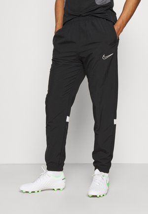 Homme portant un pantalon Nike noir, les mains dans les poches, et des crampons blancs avec des touches vertes, debout devant un fond uni.