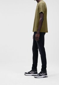 Olivgrünes Baumwoll-T-Shirt mit kurzen Ärmeln, kombiniert mit schwarzen, schmal zulaufenden Jeans und gemusterten schwarzen Sneakern. Minimalistisches Design, Seitenansicht.