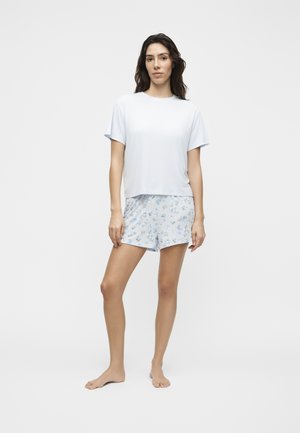 Frau steht barfuß auf weißem Hintergrund, trägt ein schlichtes weißes Kurzarmtop und hellblaue Shorts mit floralem Muster.