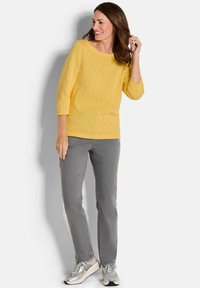 Gelber Strickpullover mit Dreiviertelärmeln, kombiniert mit grauen Hosen und silbernen Sneakern. Strukturmuster im gesamten Pullover.