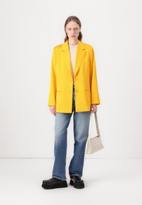 Gul oversized blazer, to knapper, skreddersydde rever, kombinert med lys blå flip-flare jeans og svarte platåklokker, holder en liten beige veske.