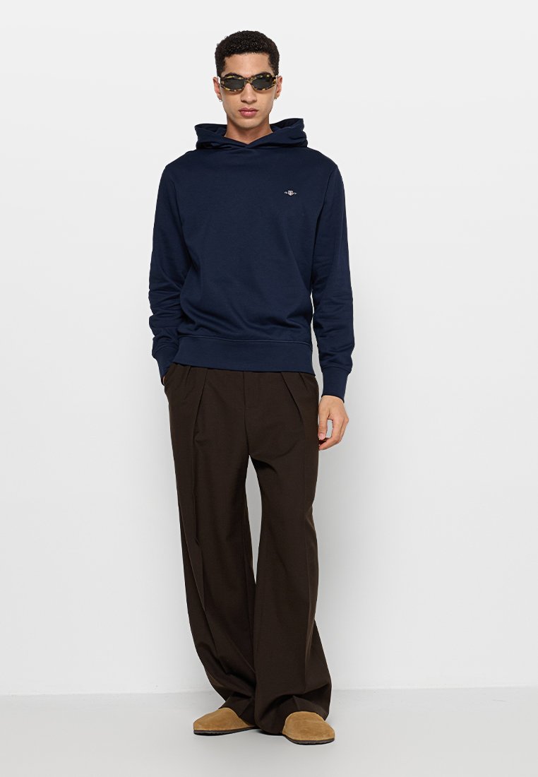 Mann trägt einen dunkelblauen Hoodie, weite braune Hosen, hellbraune Schuhe und eine Schildpatt-Sonnenbrille, stehend vor einem weißen Hintergrund.