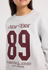 Girl wearing a light grey sweatshirt with maroon text "New York 89 Forever & Always The City of Dreams" on the front.