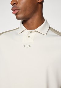 Polo shirt beige clair avec un plastron à deux boutons, arborant un logo subtil et des accents d'épaule beiges contrastants. Texture de tissu lisse.