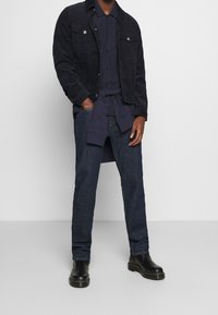 Mörka denimjeans i kombination med en marinblå knapp-up skjorta och en matchande marinblå kordjacka. Utseendet fulländas av svarta läderankelstövlar.