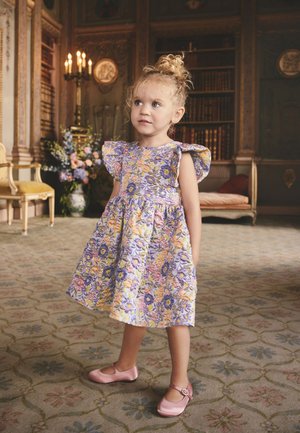 Niña joven con pelo rizado, vestida con un vestido floral y zapatos rosas, está de pie sobre una alfombra con dibujos en una habitación ornamentada con estanterías y flores.