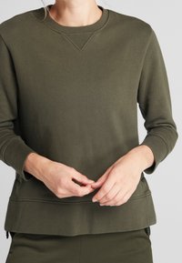 Sudadera verde oliva con cuello redondo, mangas largas y un acabado texturizado. Presenta un sutil detalle de costura en el cuello y los puños.