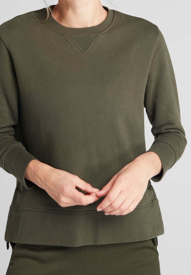 Sudadera verde oliva con cuello redondo, mangas largas y un acabado texturizado. Presenta un sutil detalle de costura en el cuello y los puños.
