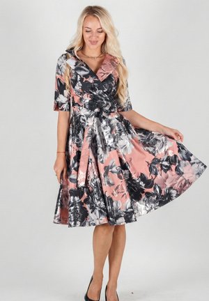 Blonde woman wearing a knee-length pink, black, and white floral dress with elbow sleeves, holding the skirt out, standing against plain background.