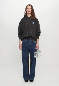 Dunkelgrauer übergroßer Hoodie mit Vordertasche, blauer Grafik, kombiniert mit hoch taillierten blauen Jeans, hält eine silberne Handtasche, schwarze Schuhe.
