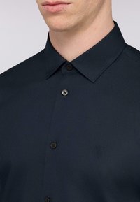 Camicia blu navy con colletto classico, dotata di chiusura a bottoni e un logo discreto. Tessuto liscio con un design pulito e minimalista.