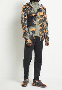 Completo sportivo composto da una giacca con motivo camouflage e accenti arancioni, un top verde chiaro, pantaloni neri e sneaker nere con texture.