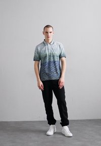 Polo krekls ar rakstu, kas piedāvā horizontālas svītras zaļā un zilā krāsā, apvienots ar melnām biksēm un baltām tenisa apavām. Vienkāršs dizains, īsas piedurknes.