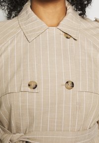 Beige gestreepte trenchcoat met een grote kraag, dubbele knoopsluiting en tailleband. Het materiaal heeft een gestructureerde afwerking.