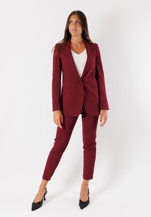 Donna in piedi con indosso un blazer bordeaux e pantaloni, una maglietta bianca e tacchi neri, davanti a uno sfondo bianco uniforme.