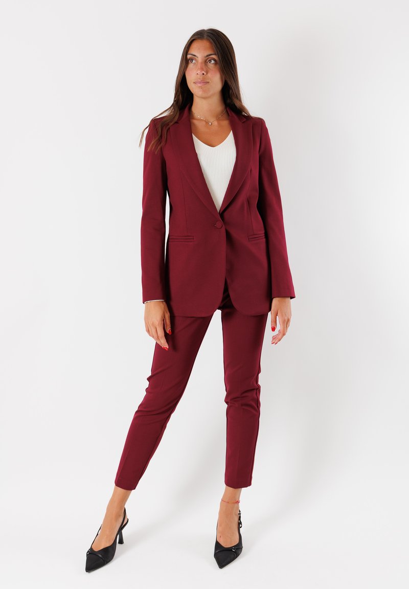 Donna in piedi con indosso un blazer bordeaux e pantaloni, una maglietta bianca e tacchi neri, davanti a uno sfondo bianco uniforme.
