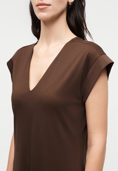 Γυναίκα που φοράει καφέ φόρεμα V-λαιμόκοψη με καπελωτά μανίκια και χρυσούς κρίκους σκουλαρίκια, εμφανίζεται από τους ώμους μέχρι τη μέση του κορμού σε λευκό φόντο.