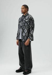 Man met gevlochten haar, gekleed in een zwart-grijs patroon jas, wit shirt, losse zwarte jeans en zwarte laarzen, staande tegen een lichte achtergrond.