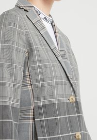 Blazer a quadri grigi con un design strutturato, caratterizzato da un colletto a revers, due bottoni e schemi a righe sovrapposti in toni di nero e beige.