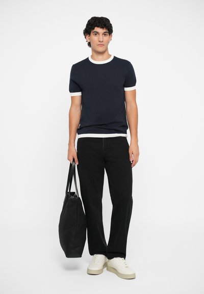 T-shirt de malha azul-marinho com colarinho e punhos brancos em canelado, emparelhado com calças pretas e ténis brancos. A segurar uma mala tote preta.