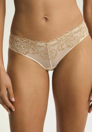 Beżowe koronkowe majtki typu hipster z kwiatowym haftem noszone przez osobę, ukazującą środkową część ciała i górną część ud na neutralnym tle.
