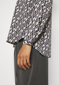 Camicia a maniche lunghe con motivo a rombi nei colori blu navy, crema e nero, con orlo tondo e tessuto morbido. Mano appoggiata su pantaloni grigi.