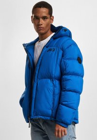 Kék puffert jacket, kapucnival, cipzáros záródással és elasztikus mandzsettával. Logo a mellkason és texturált anyag, steppelt mintázattal.