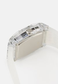 Montre rectangulaire transparente avec une couronne argentée et un bracelet en silicone transparent ; texture lisse avec un minimalisme sur le cadran de la montre.