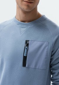 Ljusblå sweatshirt med en kontrasterande ficka med en svart dragkedja. Materialet är mjukt, med en texturerad yta och synliga sömdetaljer.