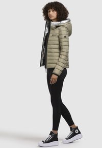 Lichtgroene pufferjack met een glanzende zilveren capuchon, ritssluiting en zwarte accenten, gecombineerd met zwarte leggings en high-top sneakers.