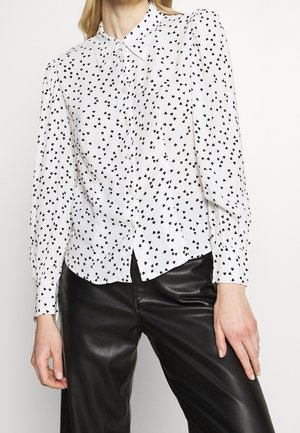 Woman wearing a white button-up blouse with black heart patterns and black leather pants, standing against a plain white background.